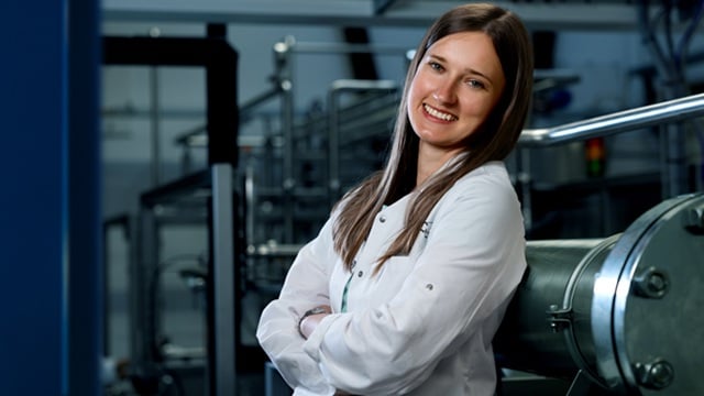  Person in a lab coat stands with arms crossed in an industrial or laboratory setting, face blurred.